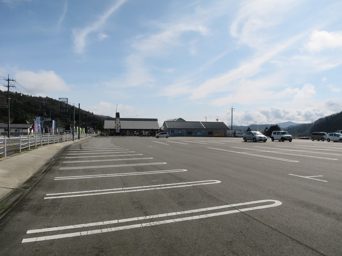 The west car park of Michi-no-Eki Kaze no Ie: rows of white-lined standard car bays lying almost entirely empty, with a dozen or so cars parked only in the row nearest the station buildings.