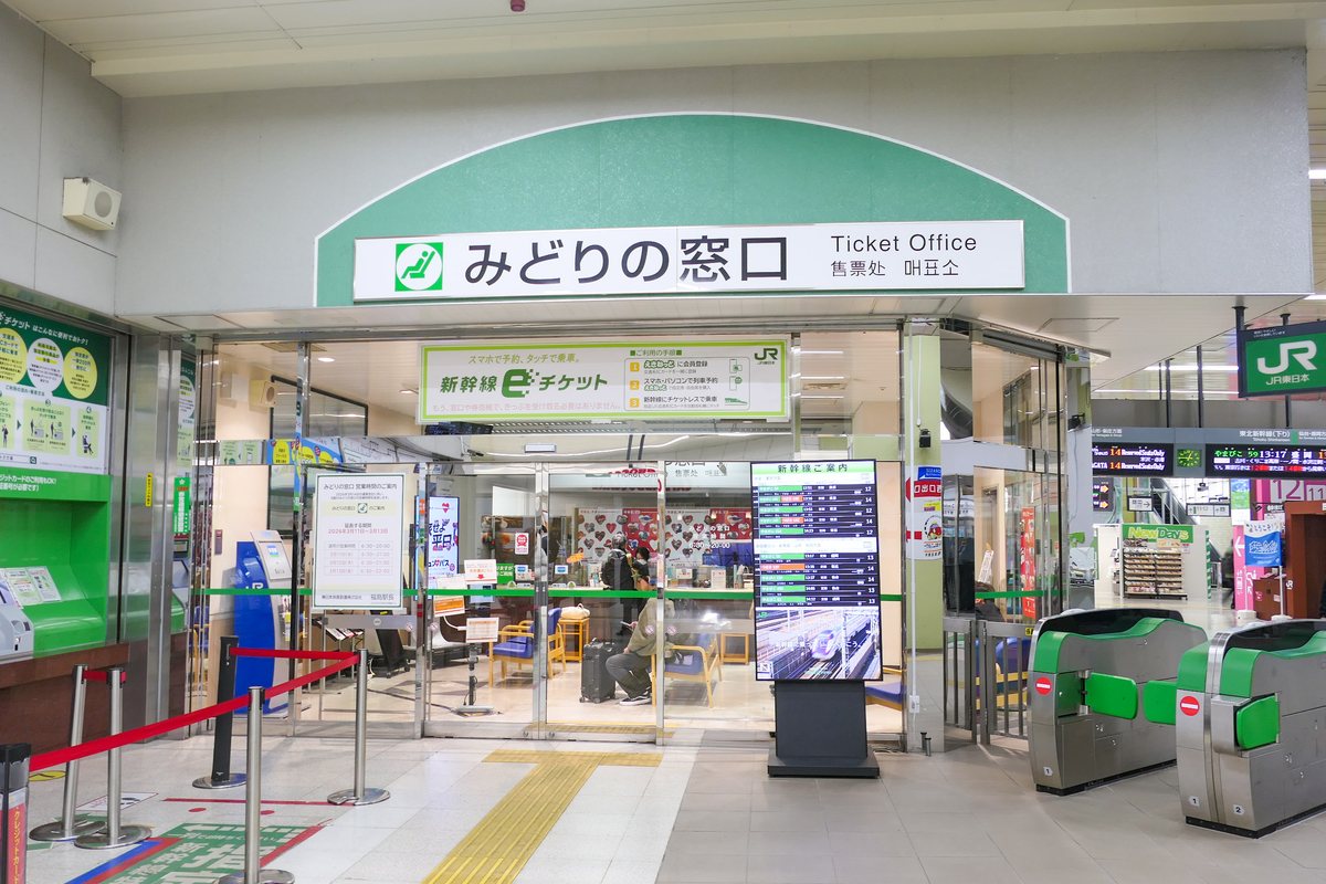 The Midori-no-madoguchi ticket office at JR East Fukushima Station, its green arched sign also reading Ticket Office, an opening-hours notice posted by the entrance and one traveller with a suitcase waiting inside