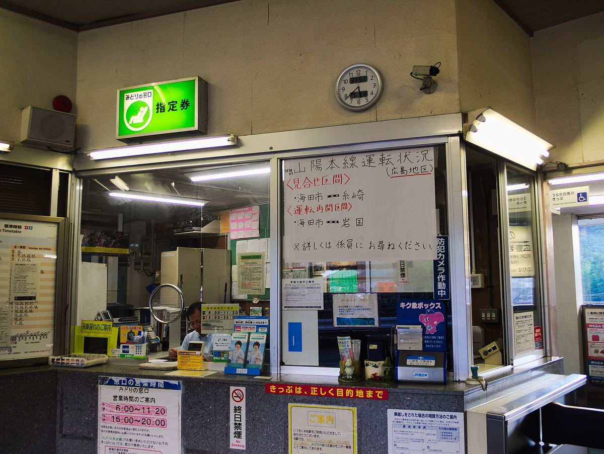 The staffed JR ticket window at Hachihommatsu Station, its green reserved-seat sign lit, with a large handwritten notice taped to the glass listing suspended and reopened sections of the Sanyo Line