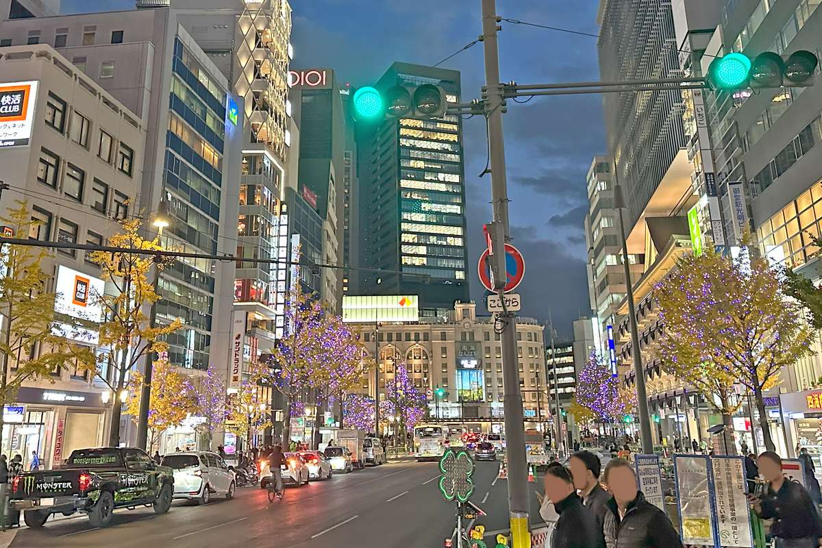 The southern stretch of Midosuji at dusk near Namba, lit street trees still carrying gold ginkgo leaves, with the Nankai Building at Namba Station straight ahead
