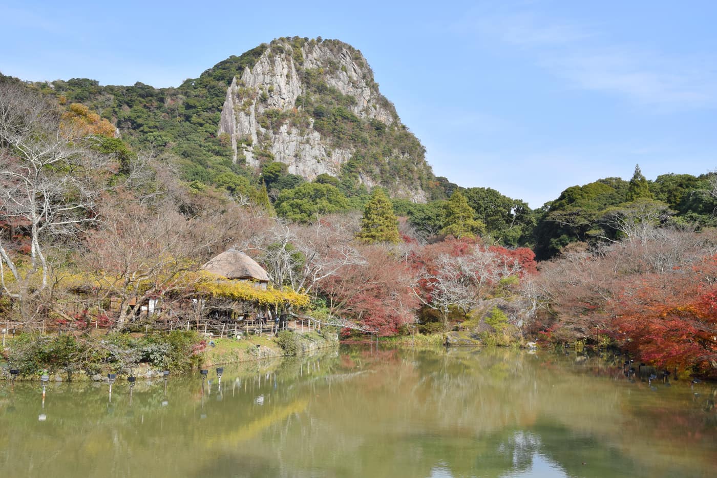 Daytime view of Mifuneyama Rakuen: the sheer rock cliff, Kagami-ike pond reflection, and autumn maples by a thatched-roof pavilion, Takeo, Saga