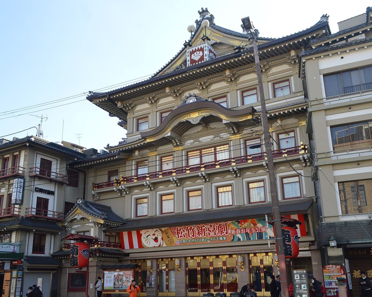 The front of Minamiza on Shijo Street in Kyoto: an undulating karahafu gable across the centre of the roof, a white-draped yagura turret above it, large red lanterns reading Minamiza hanging on either side, a printed run banner stretched below the second floor, and rows of latticed entrance doors at street level with a few small figures outside.