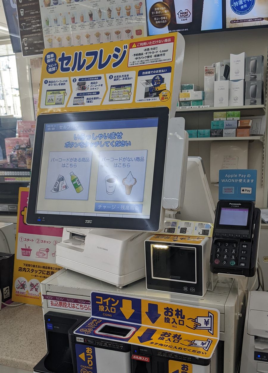 A self-checkout terminal inside a Ministop store: a yellow board above it reads self-register and shows four illustrated steps, while a red-bordered box in the top right lists the items the machine cannot handle — alcohol and tobacco, reserved goods, gift cards, Loppi, Yu-Pack drop-offs and bill collection; the screen shows a welcome prompt with two option buttons, and below sit the coin and banknote slots, with a red strip on the counter edge marking where payment-slip copies go