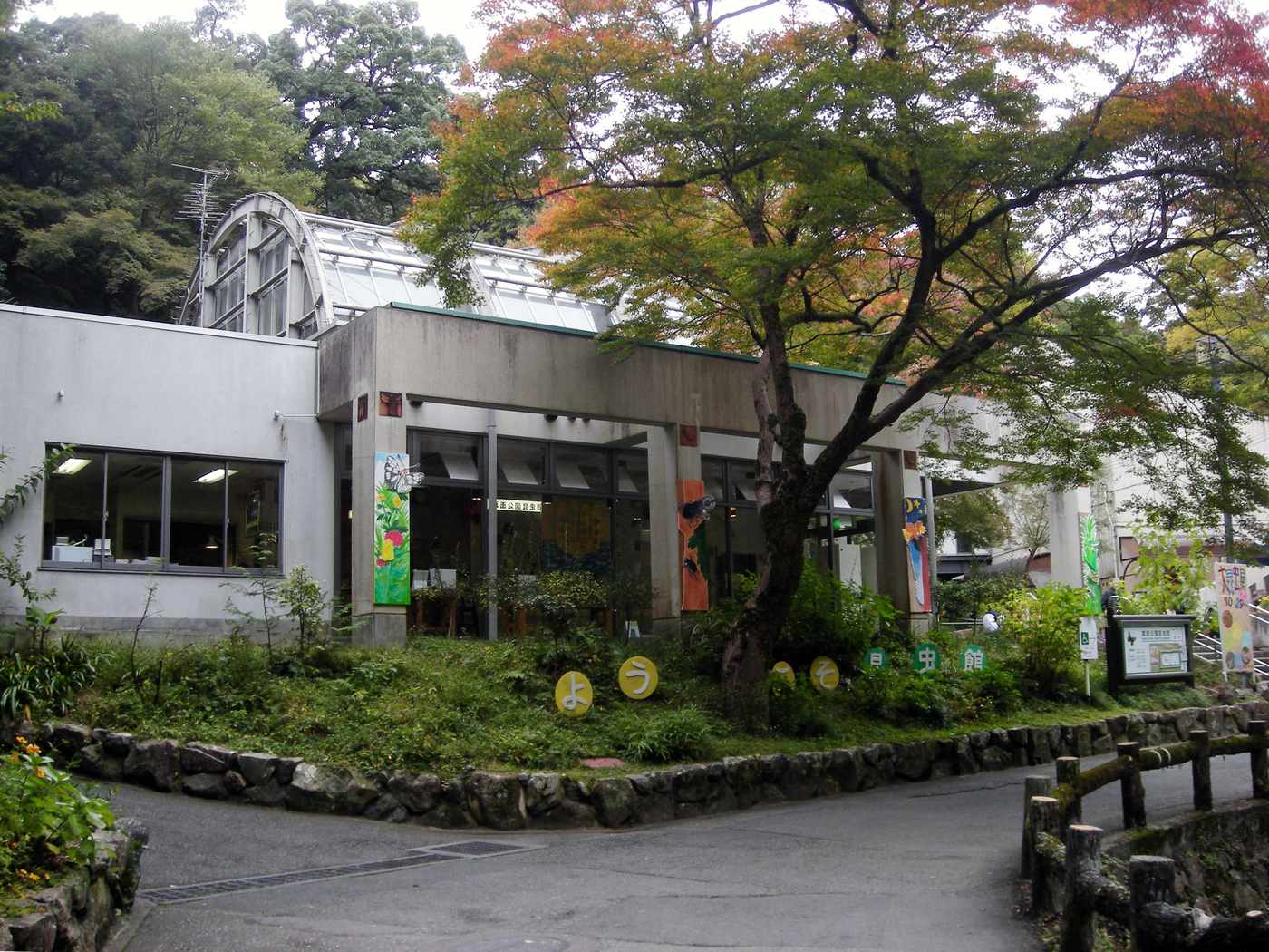 箕面公園昆蟲館外觀，混凝土館舍後方是弧形玻璃放蝶溫室，門前草坡排著平假名字牌