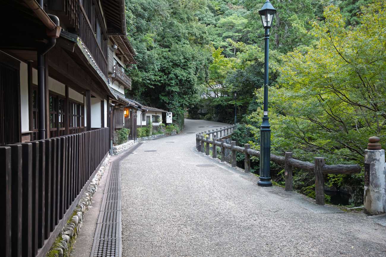 箕面滝道的鋪裝路面，平整步道沿溪谷延伸，左側是木造茶屋、右側是木柵欄與路燈，整段沒有階梯