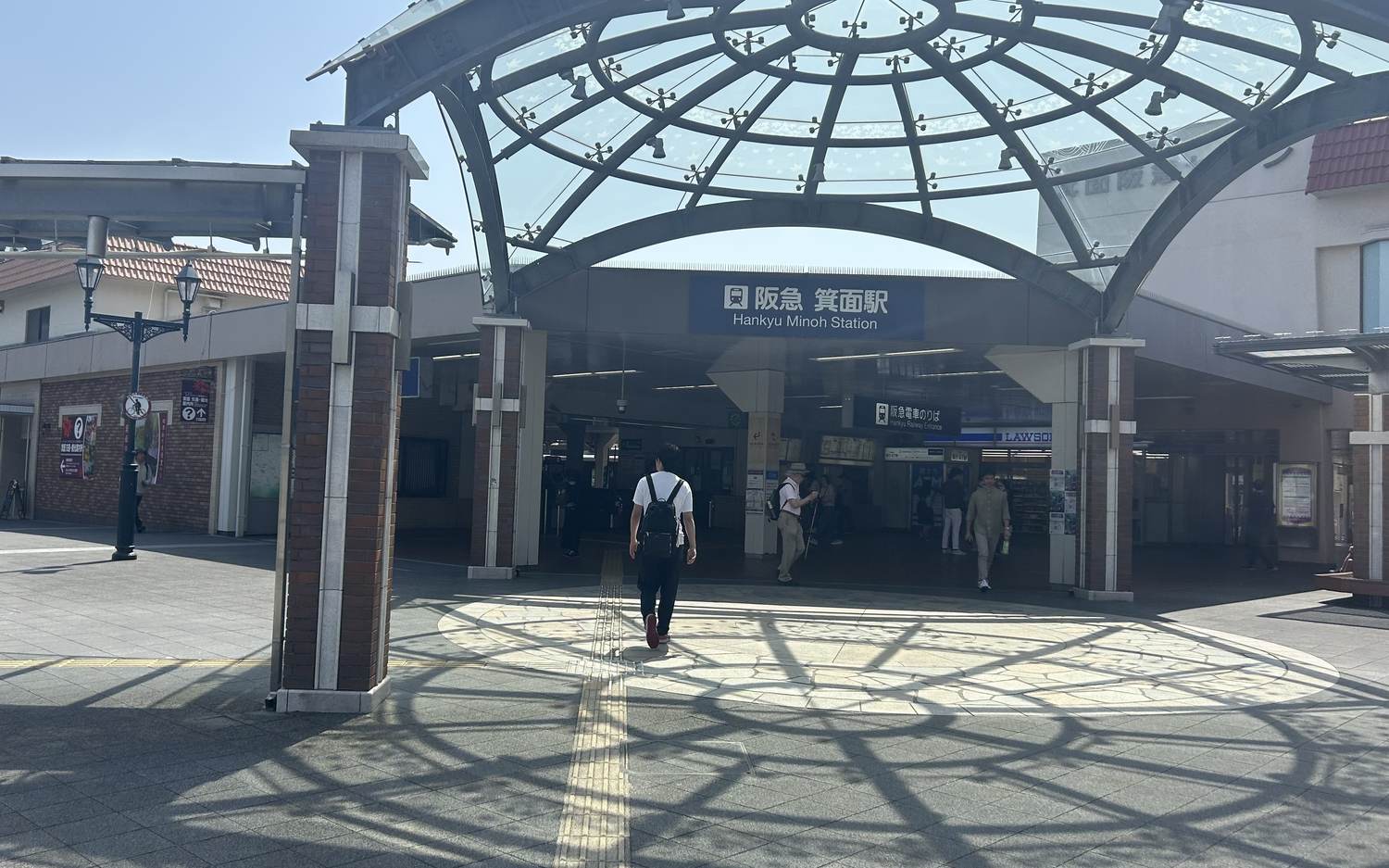 阪急箕面駅站前廣場，拱形玻璃雨棚下掛著藍底白字的「阪急 箕面駅 Hankyu Minoh Station」站名招牌，右側是便利商店與「阪急電車のりば」指標