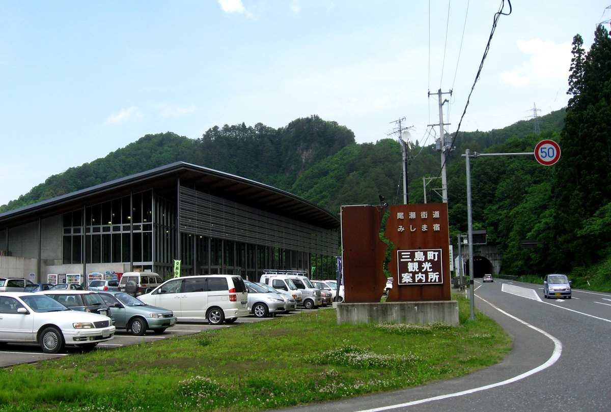 道の駅尾瀬街道みしま宿的外觀:弧形屋頂的深色建築前是停滿車的停車場,路邊一座鏽色鋼板招牌寫著「尾瀬街道 みしま宿」與「三島町観光案内所」,右後方國道盡頭是隧道口
