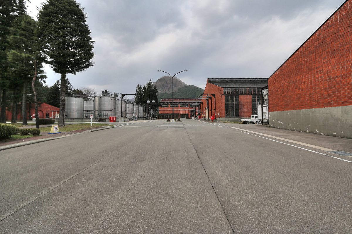 宮城峽蒸餾所廠區裡一條很寬的柏油路往前延伸,右邊是一整面紅磚牆的大廠房,遠處左邊有一排銀色不鏽鋼槽、正前方是更多紅磚建築,後面一座尖頂的山;路左側種著幾棵高大的杉樹,遠處停著一輛小貨車,陰天,路上沒有人