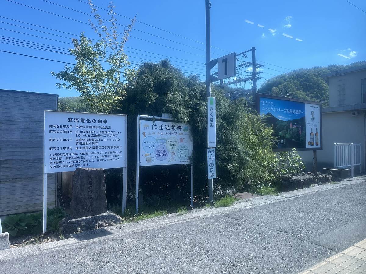 隔著車窗看作並站月台:左邊是「交流電化の由来」說明板與「作並温泉郷」旅館介紹板,中間立著「さくなみ」站名柱與「山寺・山形方面のりば」,右邊是一面寫著「ようこそ、ニッカウヰスキー宮城峡蒸溜所へ」的大型看板,畫面上有一點車窗反光