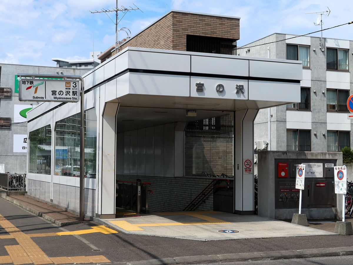 宮の沢站的地面出入口:白色方形雨庇下是往地下的樓梯,正面寫著「宮の沢」,左邊立著綠色的地下鐵站牌,牌上標著東西線的橘色圖示與「3 出入口」、「宮の沢駅 Miyanosawa Sta.」