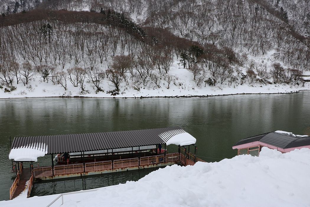 冬天的最上川草薙港：前景是積雪的河岸與有屋頂的浮動棧橋，對岸整面山坡覆著雪、只剩落葉樹的枝幹，河水是深綠色