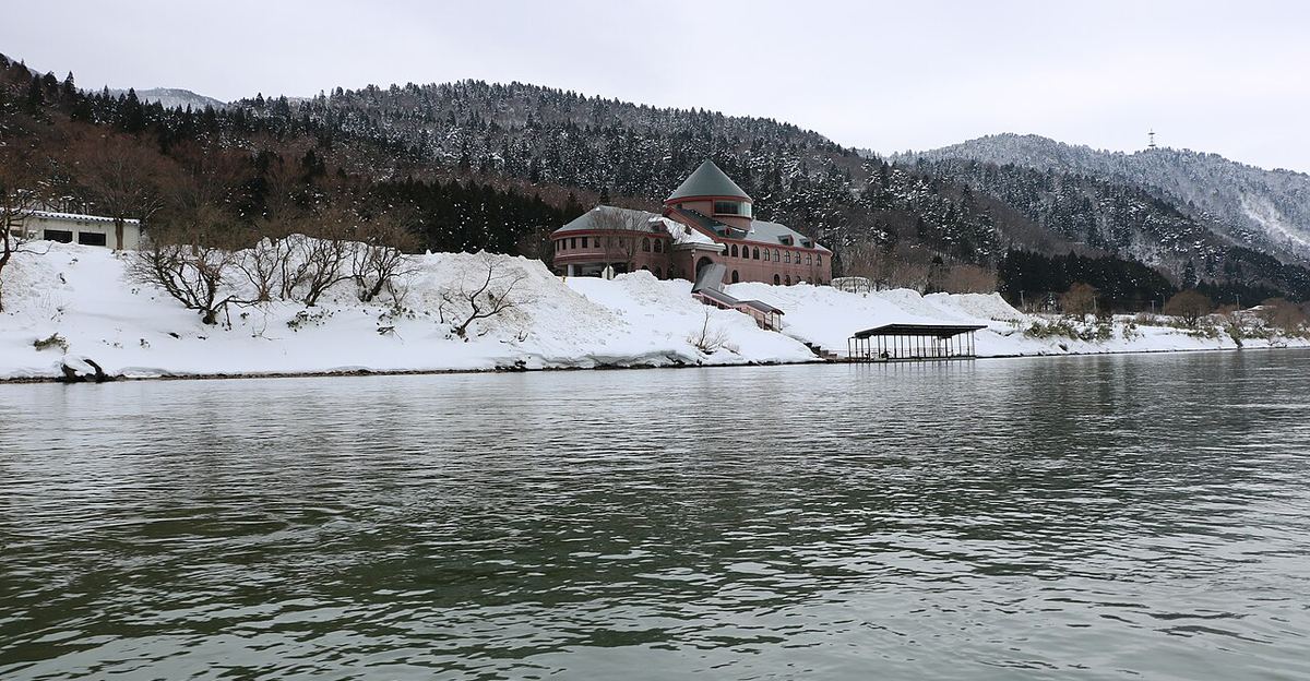從最上川河面上看草薙港：積雪的河岸上有一棟紅磚色、圓錐形綠屋頂的建物，岸邊有一座有屋頂的浮動棧橋，背後是覆雪的杉林山丘