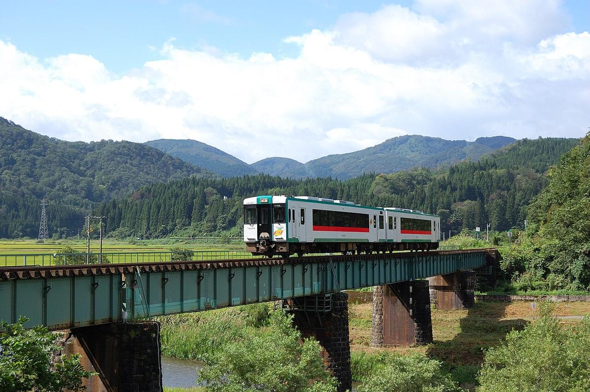 兩節編組的白底綠帶柴油列車開過一座綠色鋼梁鐵橋，橋下是小河，背景是稻田與杉林山丘