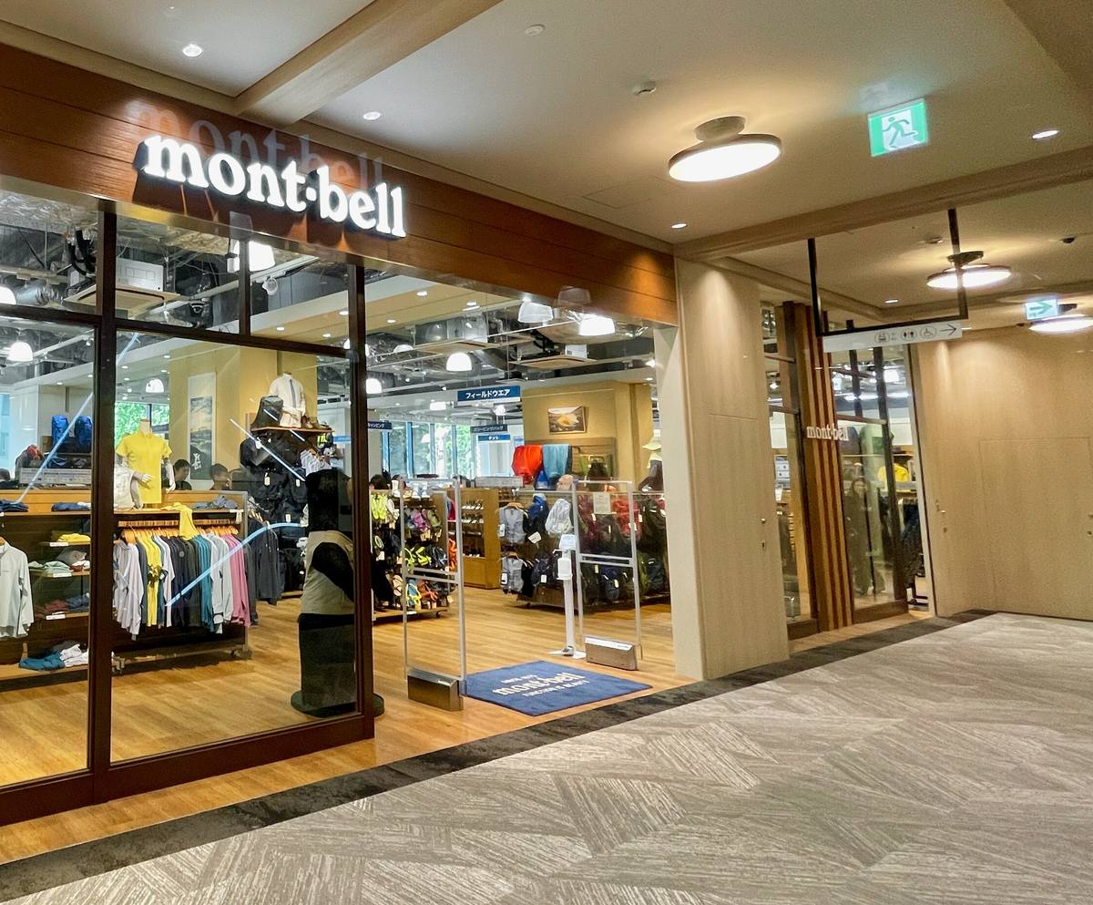 商場走廊上的 Montbell 名古屋栄店入口:木紋門楣上亮著白色 mont-bell 字樣,玻璃後面是掛著 T 恤與薄外套的衣架和人形模特兒,入口地上一塊藍色地墊,右邊是商場的米色牆面與廁所指示牌
