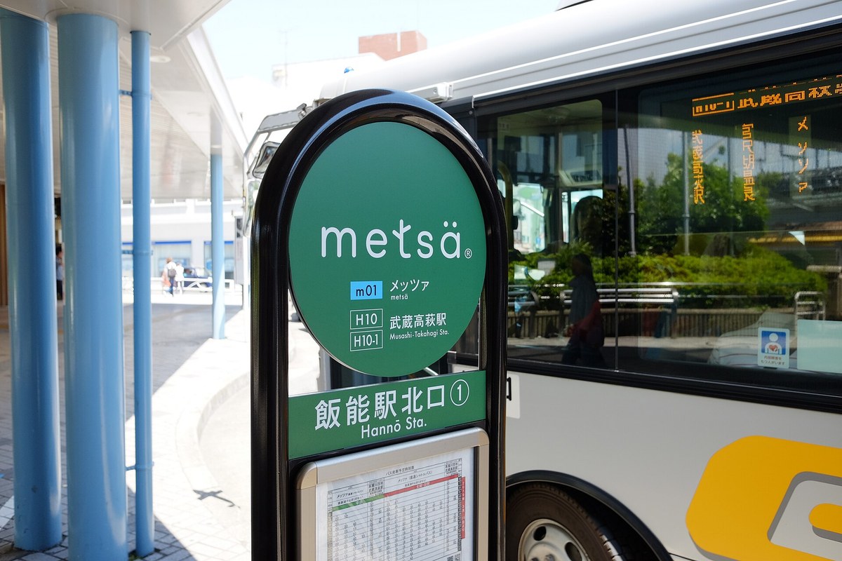 飯能站北口的巴士站牌:綠色圓牌寫 metsä、m01 メッツァ與 H10 武蔵高萩駅,下方寫「飯能駅北口①」,站牌下掛著時刻表,右邊停著一輛白色巴士,車窗上的跑馬燈寫著メッツァ