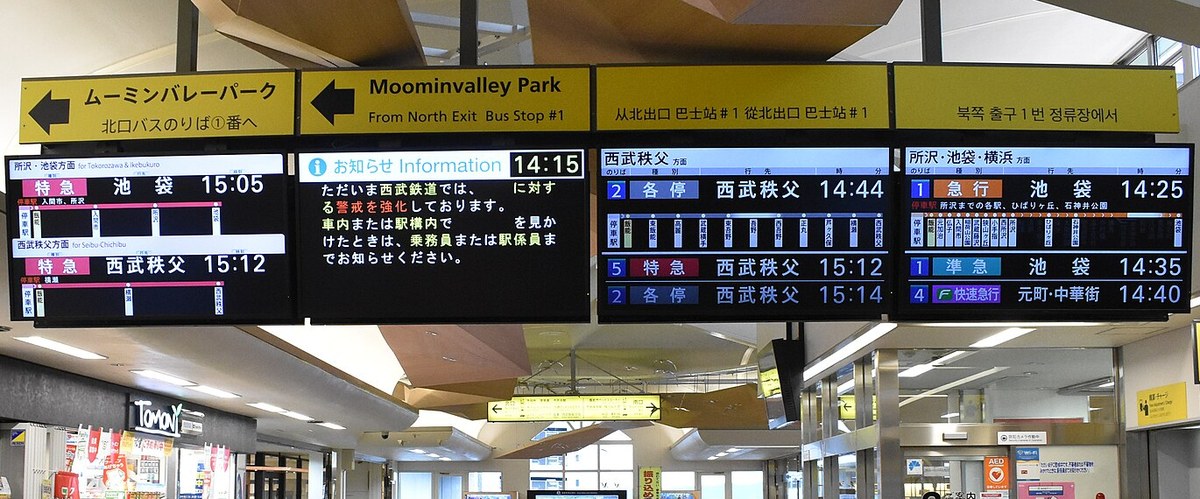 飯能站剪票口前方懸掛的黃色指標:左邊寫「ムーミンバレーパーク 北口バスのりば①番へ」與英文 Moominvalley Park From North Exit Bus Stop #1,旁邊是中文與韓文的同樣說明,下方是一排電車發車看板