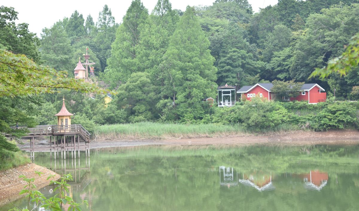 從嚕嚕米樂園內看宮沢湖:左前方湖面上有棧橋與黃色尖頂小屋,對岸樹林間是兩棟紅色木屋、一座粉紅色尖塔與一根船桅,陰天,湖面是綠色的倒影