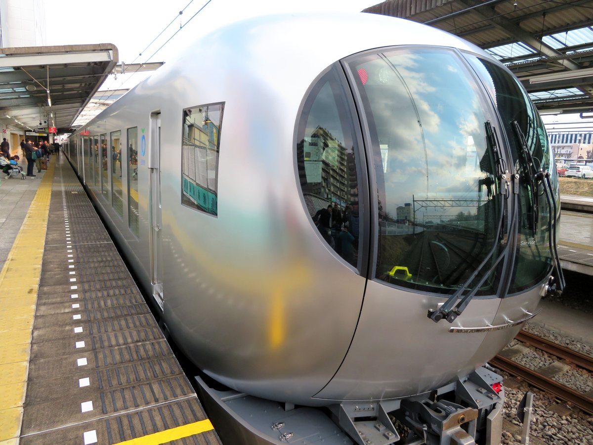 特急 Laview 停在飯能站月台:銀色車身、圓弧形的車頭與一大片球面擋風玻璃,玻璃上映著天空與大樓,左邊是黃線與月台,遠處有一些等車的旅客