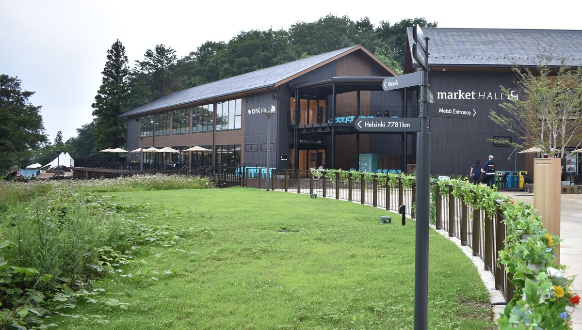 metsä village 的兩棟深灰色木造大樓:左邊外牆寫 VIKING HALL,右邊寫 market HALL,前面是一片草地與木柵欄,路標指向 Metsä Entrance 與 Helsinki 7781km,陰天