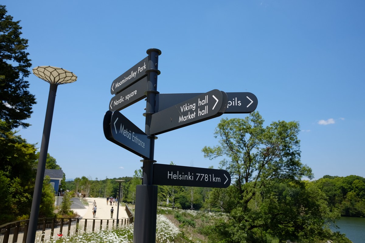 metsä village 裡的黑色路標,箭頭分別寫 Moominvalley Park、Nordic square、Metsä Entrance、Viking hall/Market hall 與 Helsinki 7781km,左邊一盞葉片造型的路燈,後方是湖畔步道與藍天