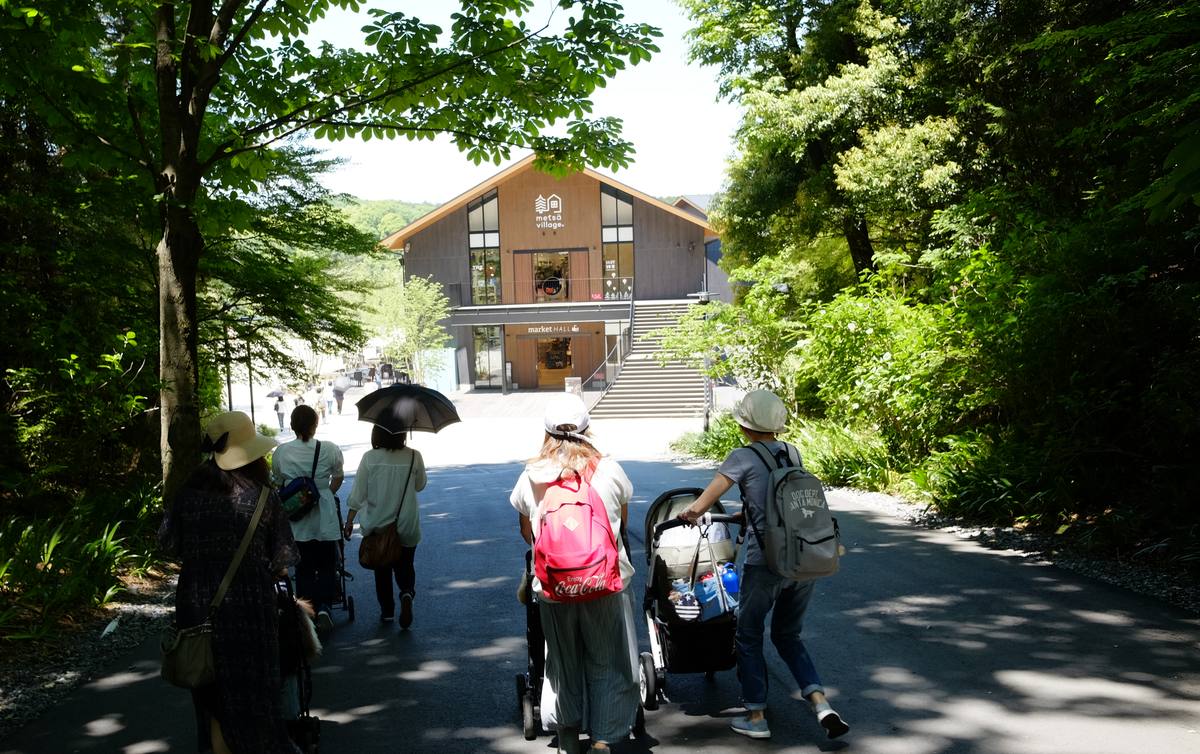 林蔭下的柏油路,幾位遊客背對鏡頭往前走,其中兩人推著嬰兒車,路的盡頭是一棟木造山形屋頂建築,外牆寫 metsä village 與 market HALL,旁邊有一道長樓梯
