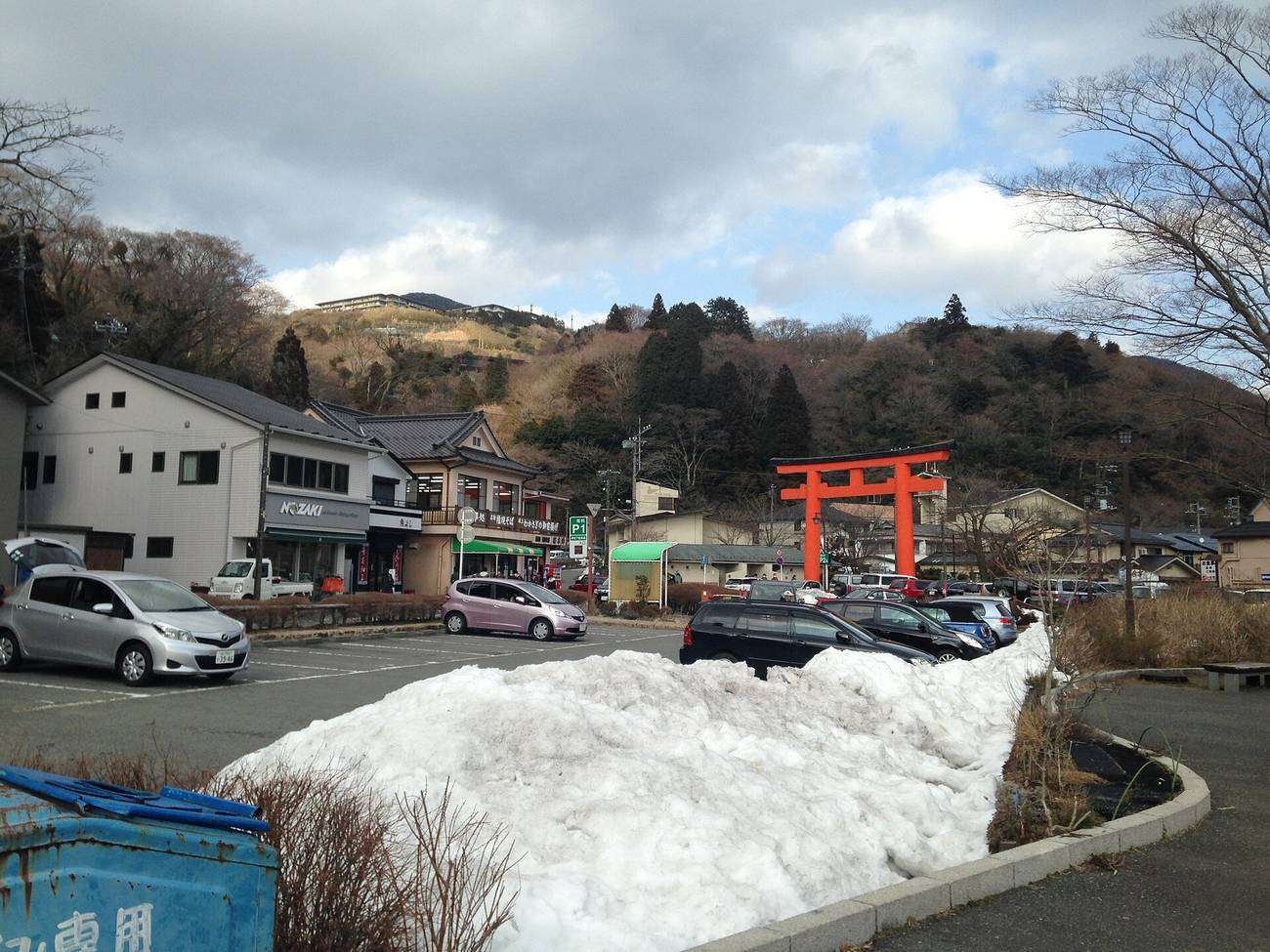A winter roadside scene at Motohakone: a vermilion torii standing at the treeline on the far side of the road, an asphalt car park full of parked cars in the foreground, a large heap of ploughed snow piled in the middle of the frame, a two-storey shop and soba restaurant on the left with a green P parking sign at the door, and bare brown hillside under an overcast sky behind