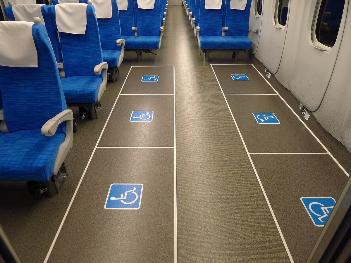 Interior of a refurbished Tokaido Shinkansen N700S car with three white-outlined floor bays on each side of the aisle, six in all, each marked with a blue wheelchair symbol; the window-side row has metal tie-down anchors and small instruction plates at the base of the cabin wall, the other row runs alongside a bank of blue seats