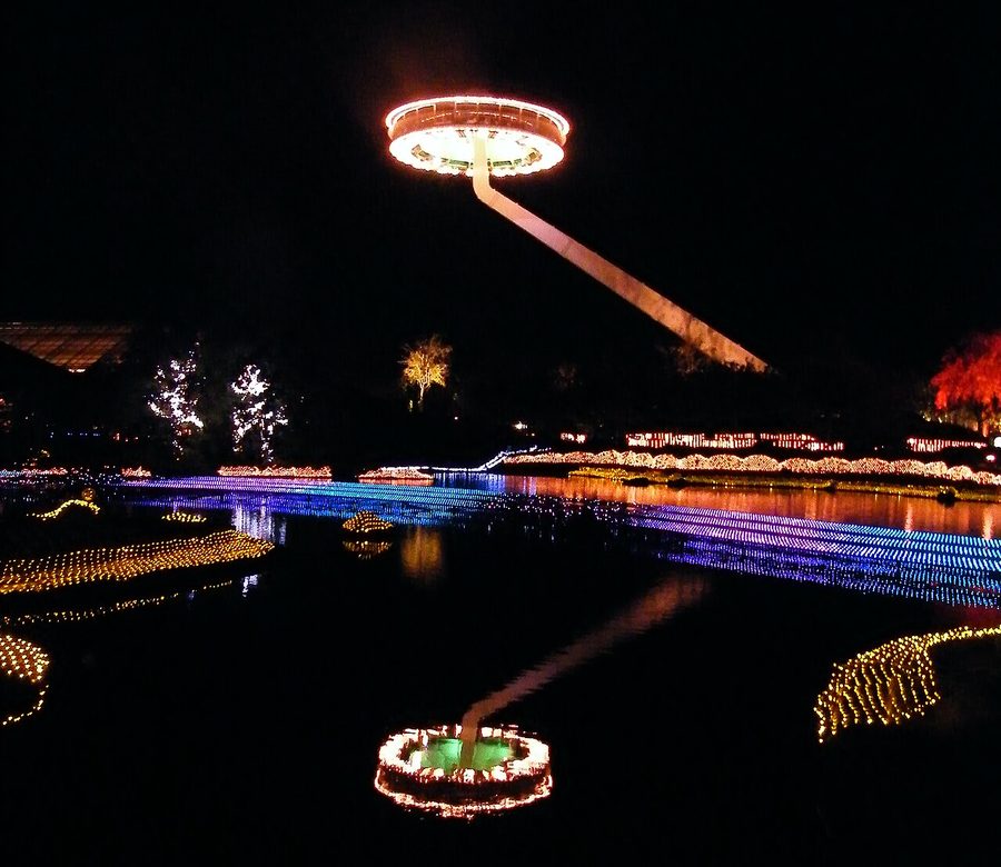 The pond at Nabana no Sato at night: a disc-shaped observation deck glowing orange is held in mid-air on a slanted arm, its reflection on the black water, with blue-purple light strips across the pond and yellow and red lights on the banks