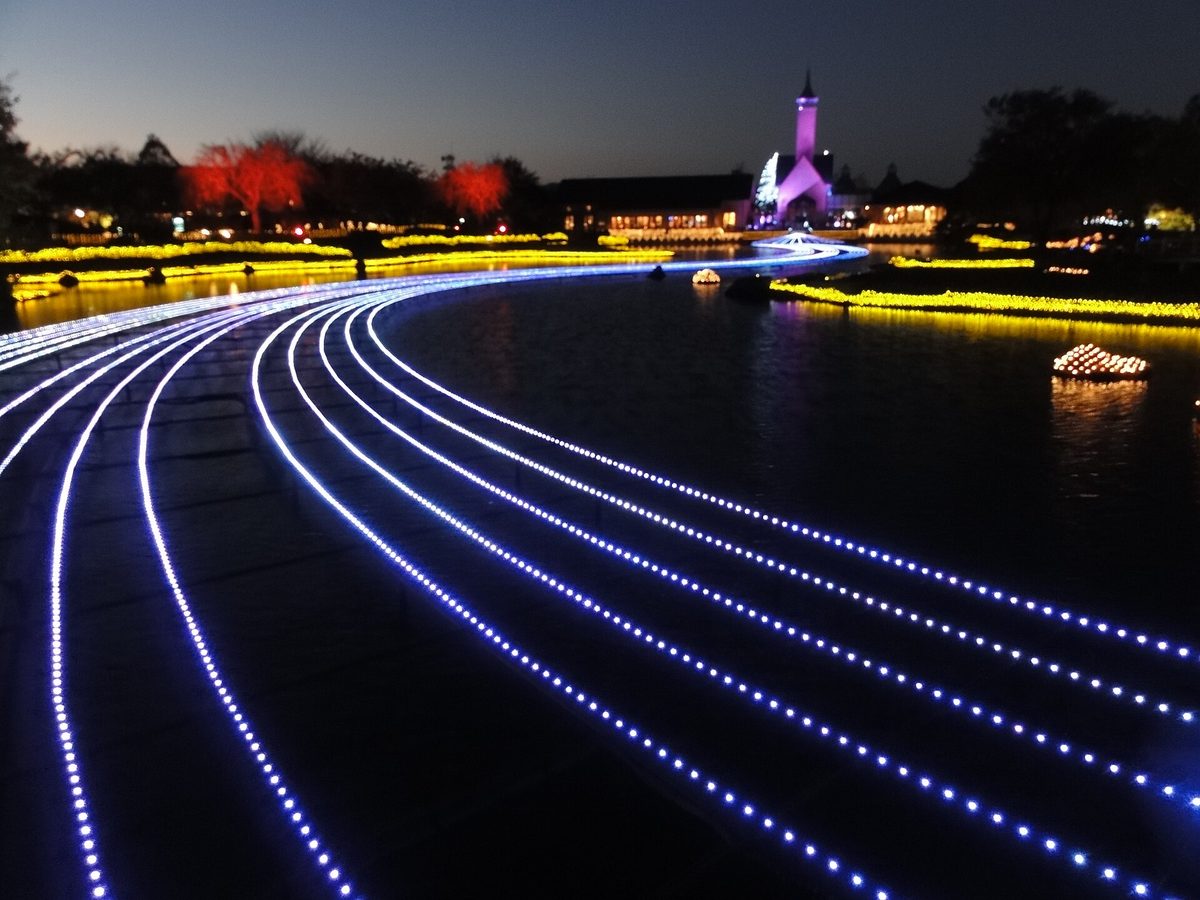 Early evening with the sky still deep blue: blue-white LED strips curve across the pond at Nabana no Sato toward a spired chapel lit in purple, with yellow light strips and two red-lit trees on the bank