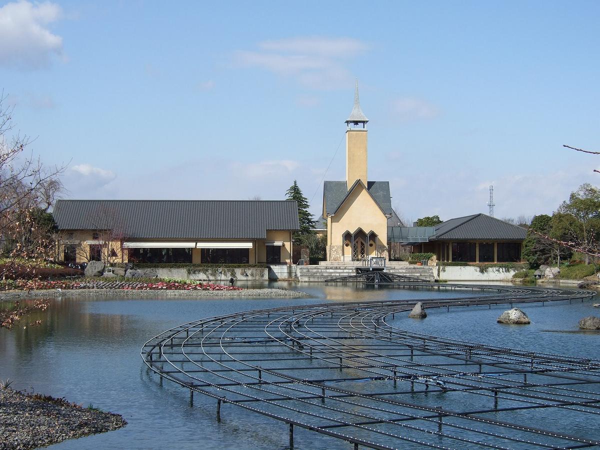 The pond at Nabana no Sato in daylight: rows of curved black metal frames stand in the water, with a cream-coloured spired chapel and grey-roofed low buildings behind, under a clear sky