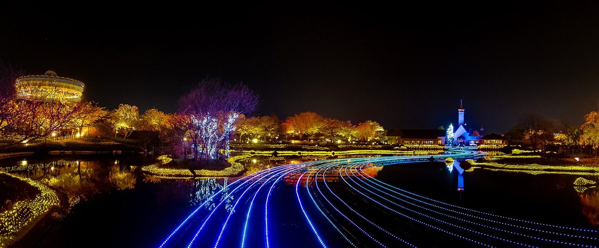 Night panorama of the pond at Nabana no Sato: blue-white LED strips curve across the water toward a spired chapel lit in blue on the far bank, with trees lit yellow and purple along the shore and a round building in gold lights on the left
