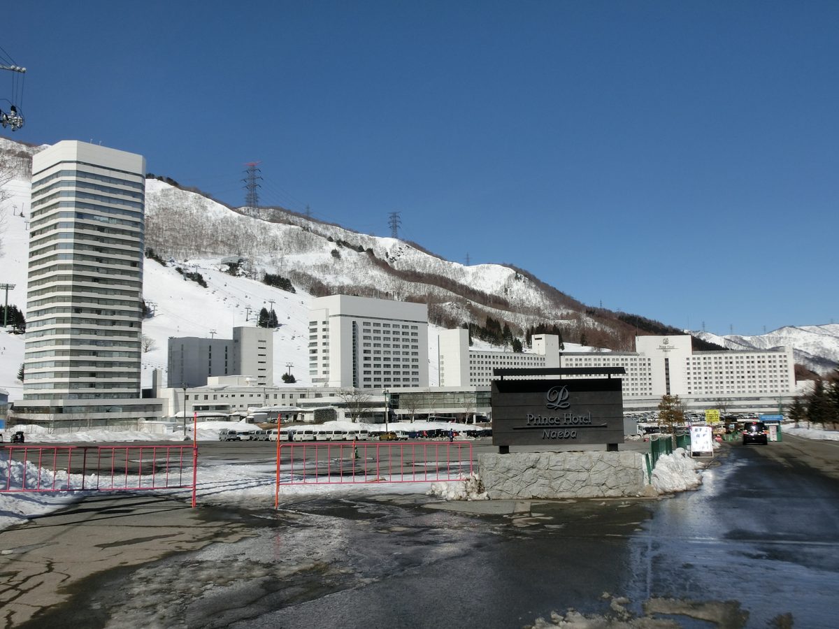 苗場王子飯店入口一帶:前景是融雪潮濕的停車場和粉紅色柵欄,右邊立著深色的「Prince Hotel Naeba」招牌,後方是一棟高塔樓和一整排相連的白色飯店大樓,再後面是有吊椅纜車和電塔的雪山
