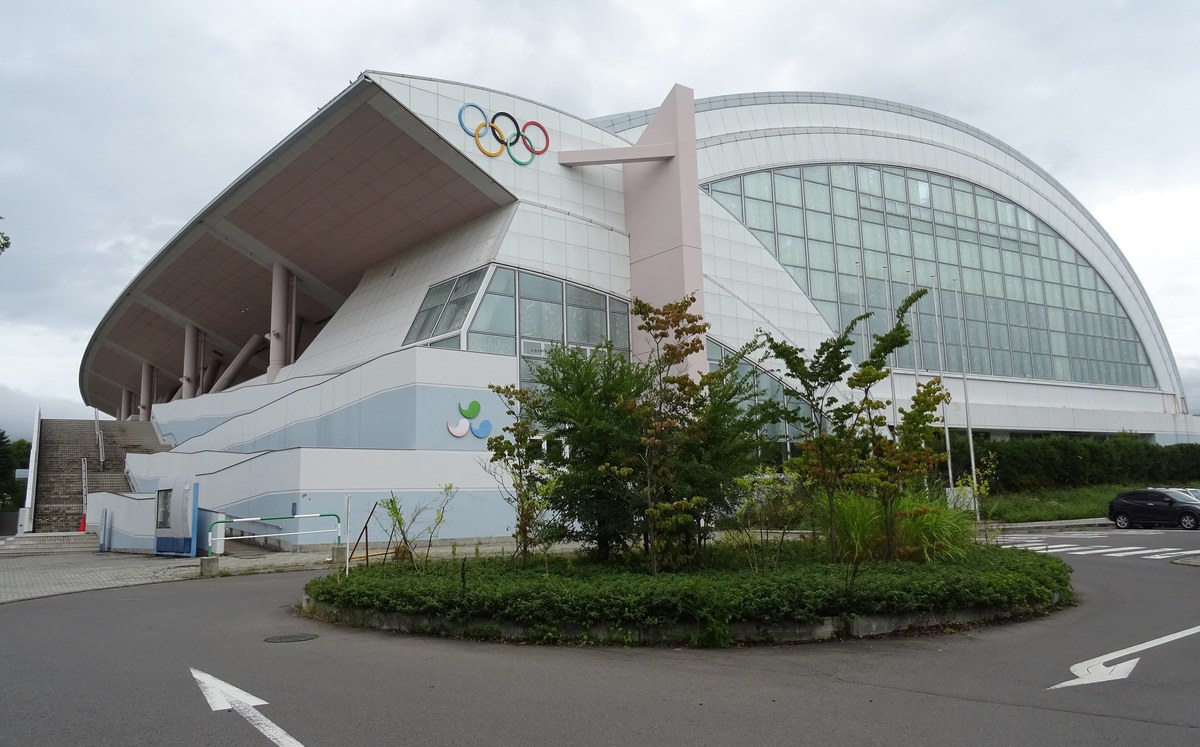 長野運動公園裡的アクアウイング:白色弧形大屋頂的室內泳池館,牆上掛著奧運五環,前方是種著小樹的圓環車道