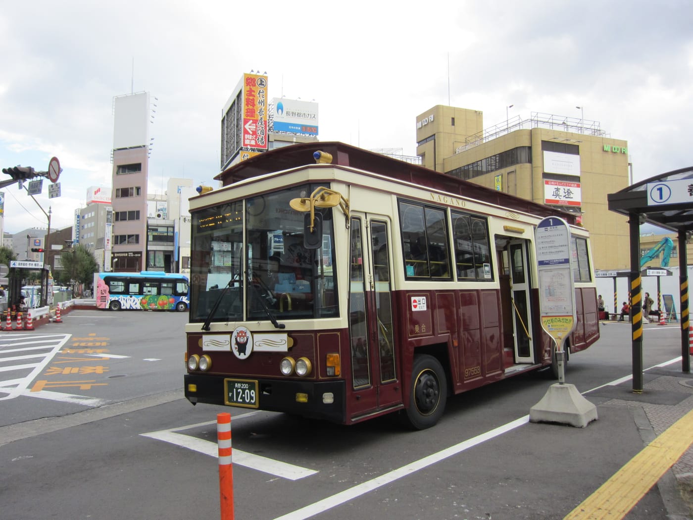 A small retro-styled bus in deep maroon and cream parked at a boarding bay in front of Nagano Station, NAGANO lettered above the windscreen and a round white mascot badge on the front, with a circular stop sign marked with a large numeral 1 and a timetable beside it