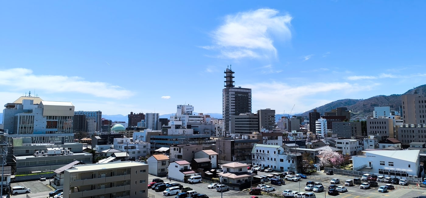 An elevated view over central Nagano in April: low houses and offices spreading to the foot of the hills, a dark high-rise topped with a white lattice mast in the centre, wooded slopes on the right, and one cherry tree in bloom beside a car park at lower right