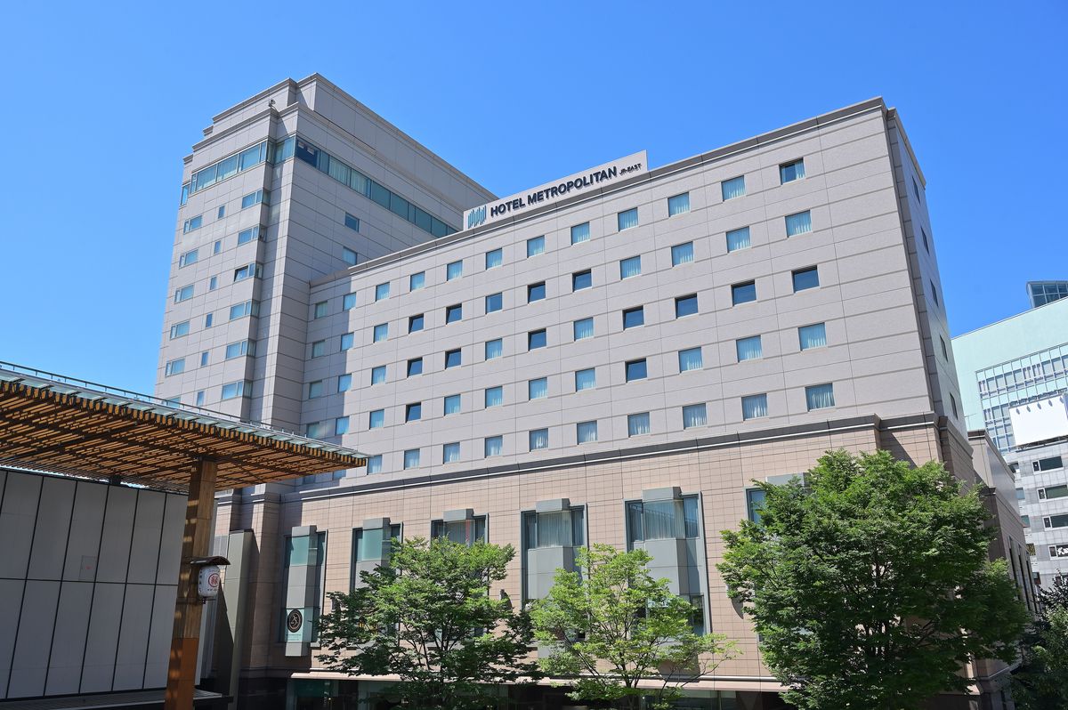晴天下的ホテルメトロポリタン長野:米灰色的高樓,屋頂招牌寫著 HOTEL METROPOLITAN JR-EAST,前方一排綠樹,左邊是長野站善光寺口的木格頂棚