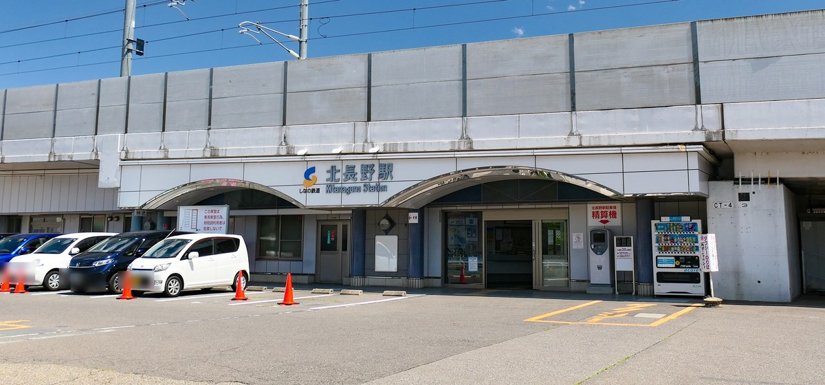 しなの鉄道北長野站的站房:高架橋下一排白色拱形入口,正中央藍字站名「北長野駅 Kitanagano Station」,前方停車場停著幾輛車