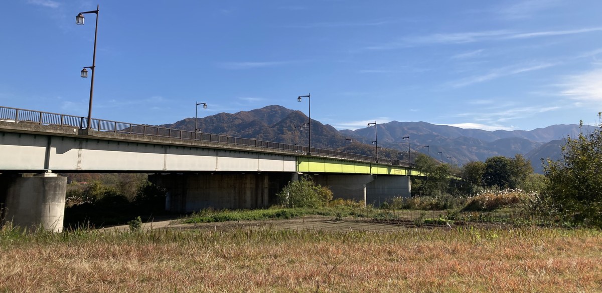 從千曲川河灘仰望松代大橋:長長的灰白色橋身架在水泥橋墩上,中段漆成黃綠色,橋上一排路燈,背後是秋天轉紅的山,前景是枯黃的草地