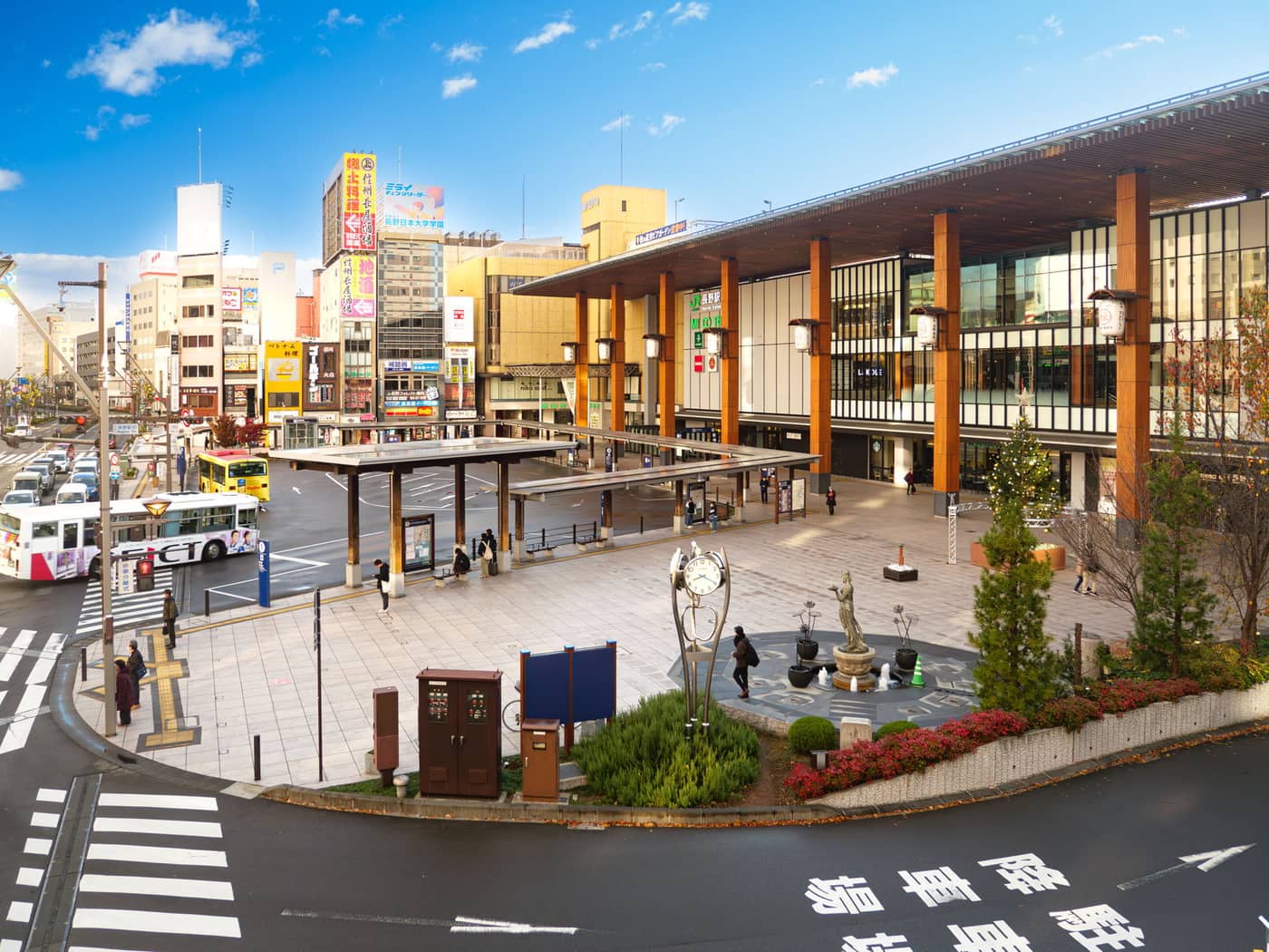 The station plaza at the Zenkoji exit of Nagano Station: a glass frontage fronted by a row of thick wooden pillars and hanging lanterns on the right, a covered bus boarding island and rotary roadway in the centre, city buildings hung with vertical signboards on the left, and a tower clock and bronze statue in the middle of the square