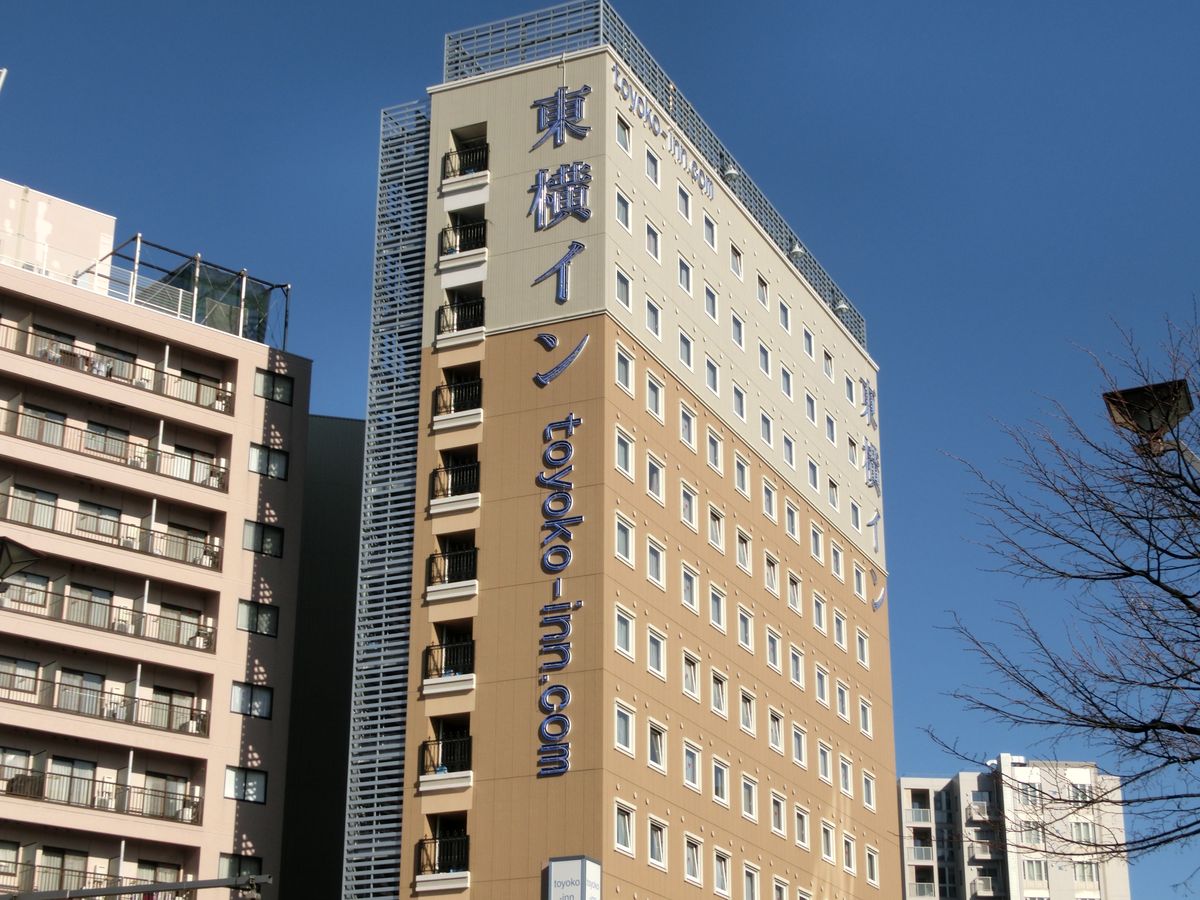 藍天下的東横イン長野駅善光寺口:細長的米黃色商務飯店大樓,牆面直書藍色「東横イン」與 toyoko-inn.com,左邊是另一棟公寓大樓