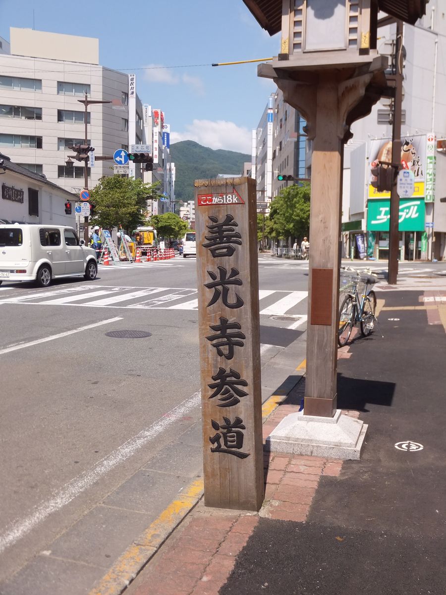 長野市中央通路邊一根木頭標柱,直書「善光寺参道」,頂端紅色牌子寫「ここから1.8k」,背後是十字路口與商業大樓
