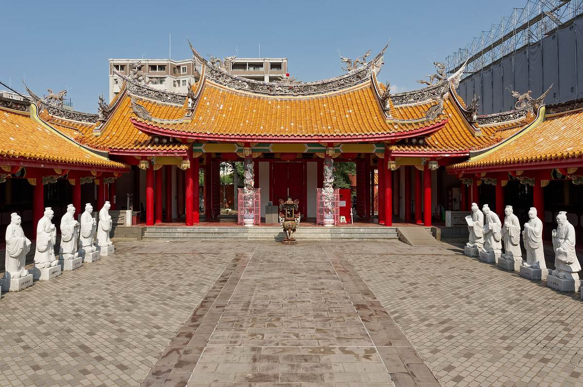 The courtyard of the Nagasaki Confucius Shrine: the main hall behind orange glazed roof tiles and vermilion columns, dragons and mythical beasts along the ridge, and two facing rows of white stone statues lining a paved central path that ends at a bronze incense burner