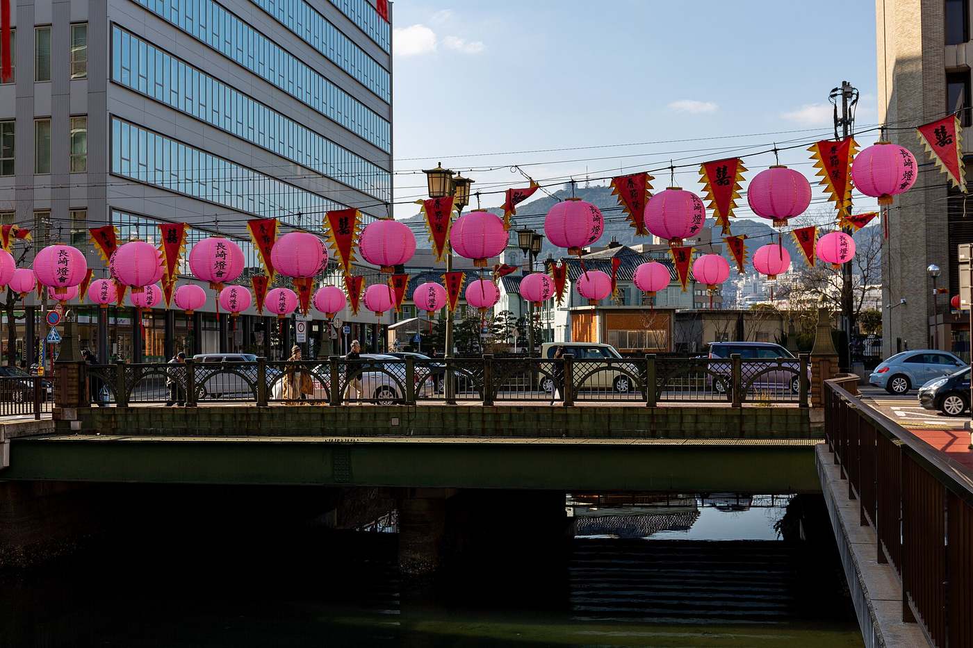 A daytime street scene in Nagasaki: a long line of pink round lanterns reading Nagasaki Lantern Festival and red-and-yellow pennants strung above the railing of a bridge over a narrow waterway, with a glass-fronted office block and a distant ridgeline under a blue sky, and cars and pedestrians crossing