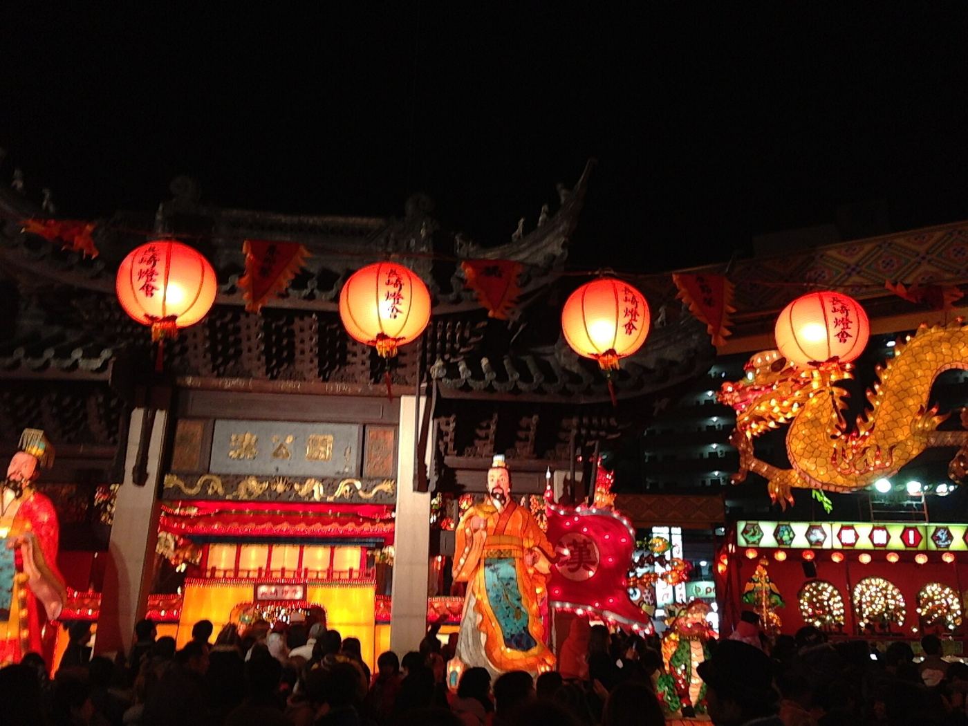 The Minato Park venue at night: four large red lanterns reading Nagasaki Lantern Festival hang from a Chinese-style gate whose plaque reads Minato Park, a golden dragon lantern curls in from the right, a robed figure lantern holding a banner stands in the centre, and a dense crowd fills the foreground
