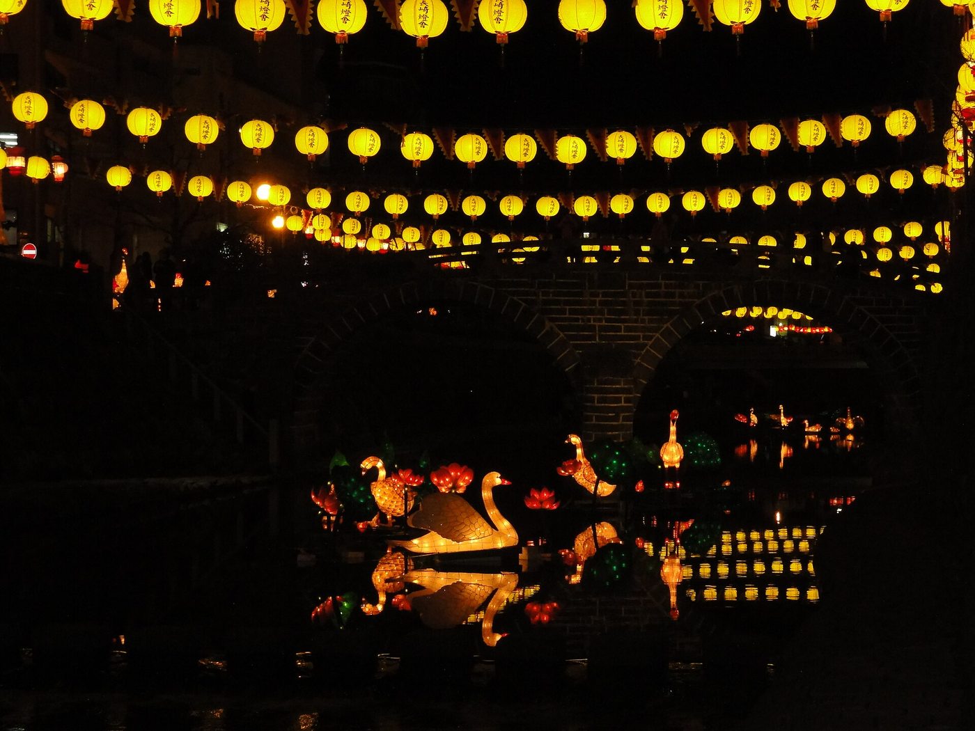 Meganebashi at night: three rows of yellow round lanterns strung above the Nakashima River, the stone arch of the bridge completing a full circle with its own reflection, and white swan and red lotus lantern sculptures floating on the water and mirrored in it