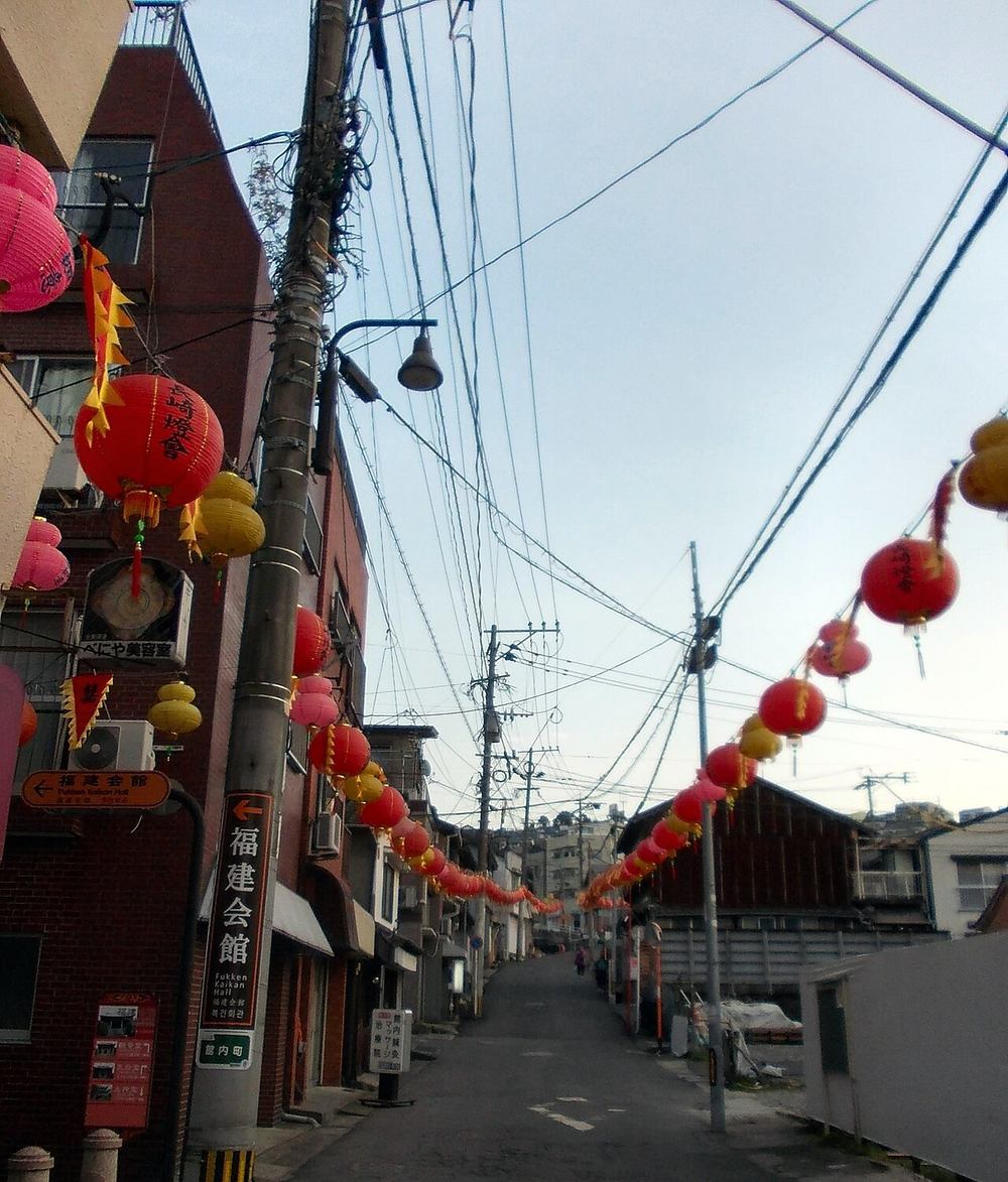 A narrow sloping lane in the Tojin Yashiki quarter: red-brick apartment blocks and older timber houses on either side, two strings of red, pink and yellow paper lanterns and red-and-yellow pennants slung across a sky full of overhead cables, a green signpost reading Fukken Kaikan Hall on the left, and a single pedestrian at the far end