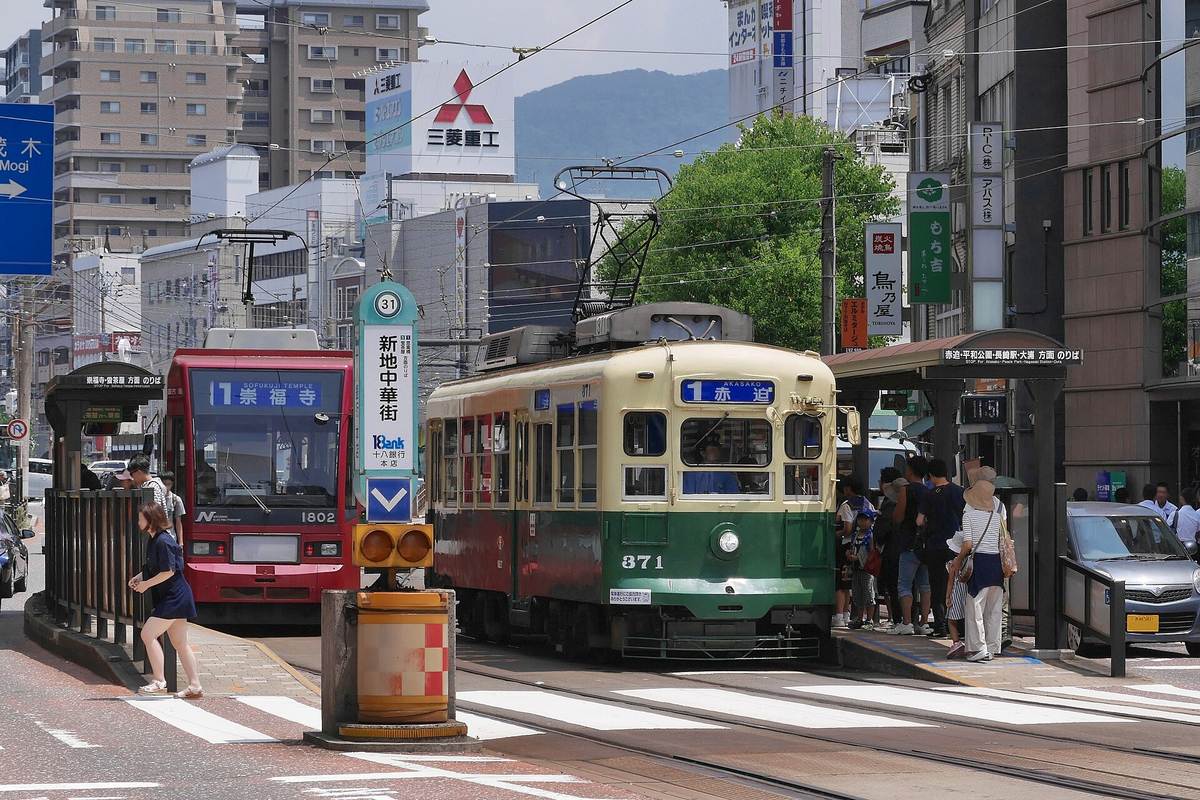 The Shinchi Chinatown tram stop in Nagasaki: an older green-and-cream tram waiting beside the island platform with a destination blind reading 1 Akasako, a red bus alongside it, a stop pillar numbered 31 and labelled Shinchi Chinatown, and passengers waiting on the platform