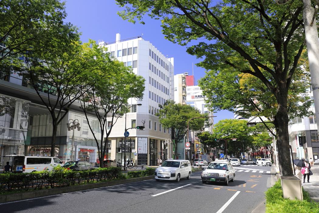 名古屋榮的大津通:兩旁是綠葉行道樹,左側是玻璃帷幕的商業大樓與藥妝店,中間路口有白色辦公大樓,路上有白色計程車與廂型車,晴天
