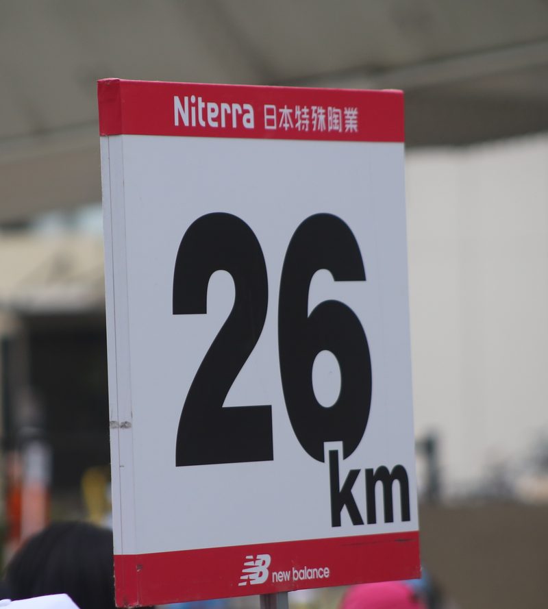 路邊立著的 26 km 距離牌:白底黑色大字「26 km」,上方紅條印 Niterra 日本特殊陶業,下方紅條印 New Balance 標誌,背景模糊