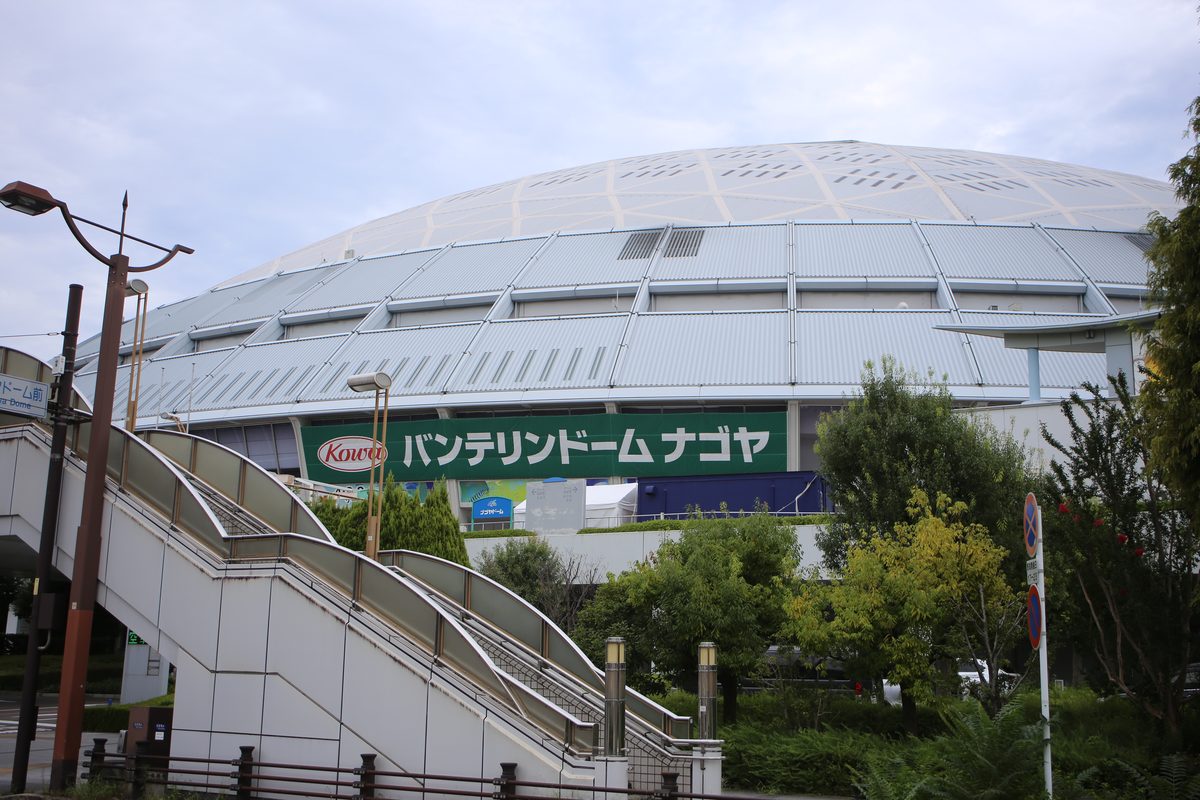 名古屋巨蛋外觀:銀白色的圓頂,牆面掛著綠底白字的「Kowa バンテリンドーム ナゴヤ」大招牌,前景是通往二樓步道的白色階梯與樹叢